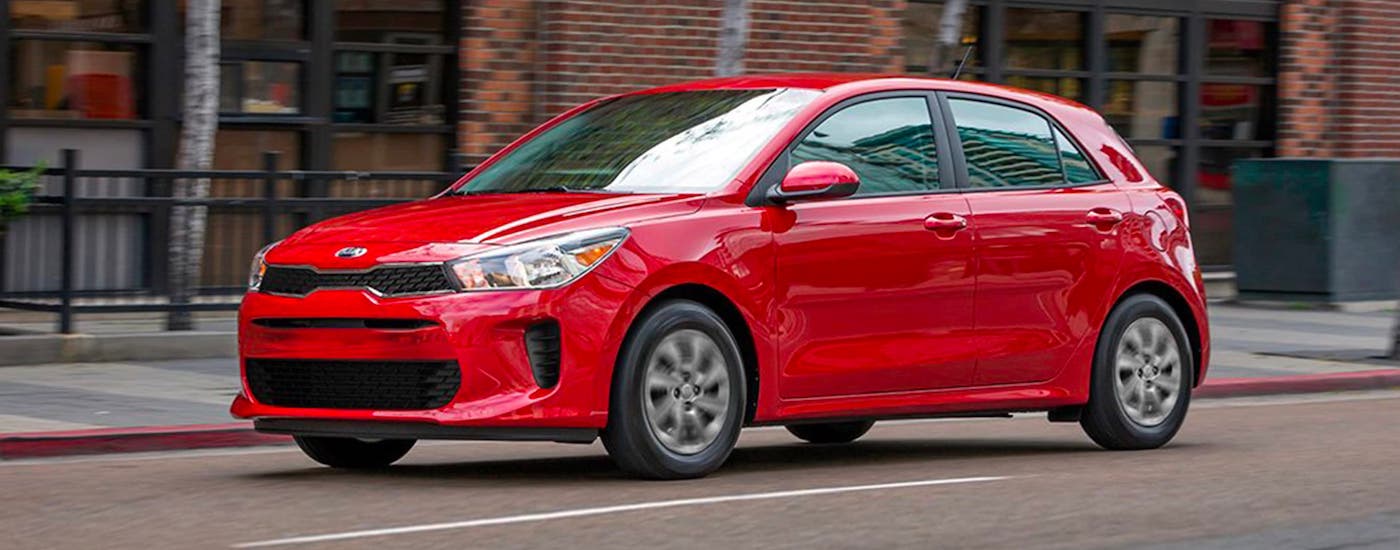 A red 2020 Kia Rio5 is driving past a red brick building.