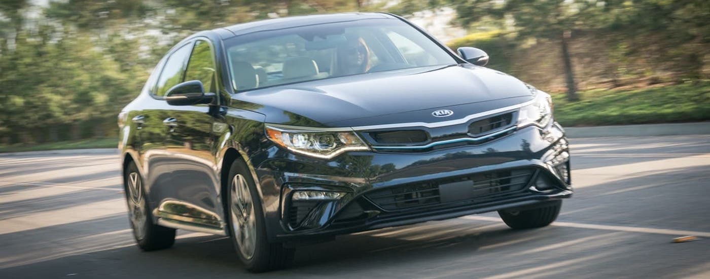 A black 2020 Kia Optima Hybrid is driving on a a street in front of trees.
