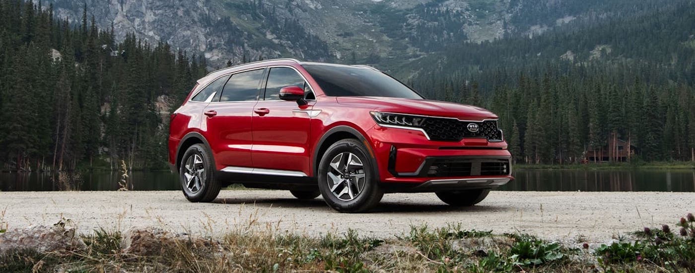 A red 2021 Kia Sorento Hybrid is parked in front of evergreen trees and a lake.